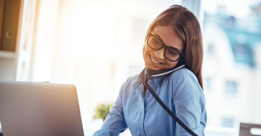 Smiling woman wearing glasses, talking on the phone while working on a laptop