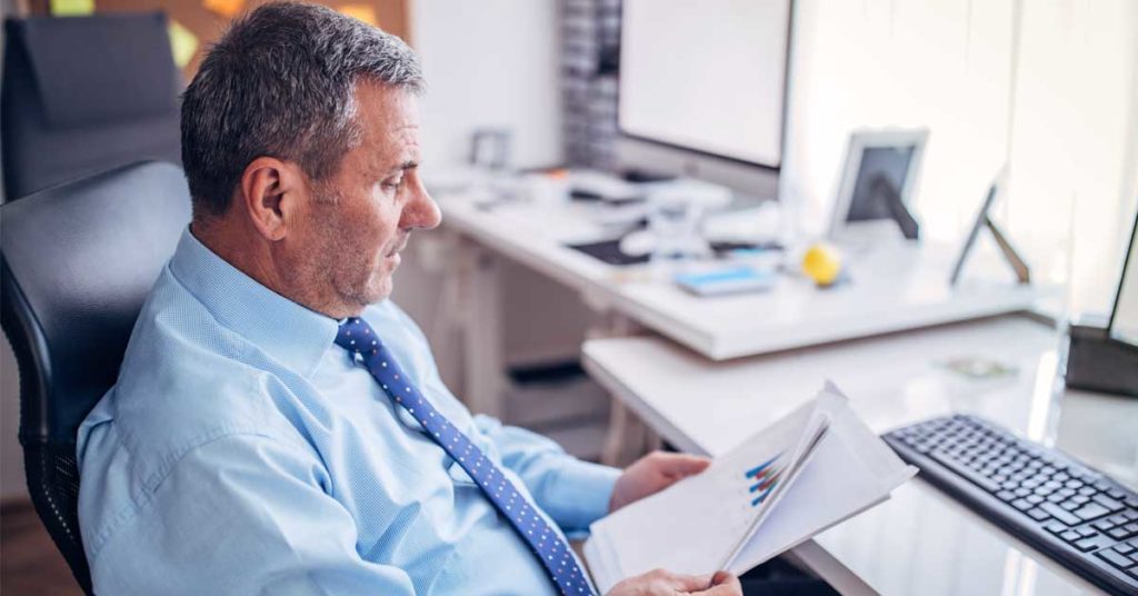 Man in a blue shirt and tie sitting at a desk, looking at papers with charts