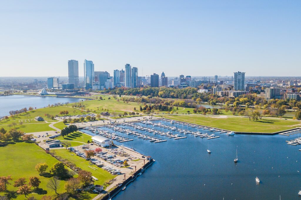 Aerial view of a marina with boats docked, a large green park, and a city skyline in the background under a clear blue sky