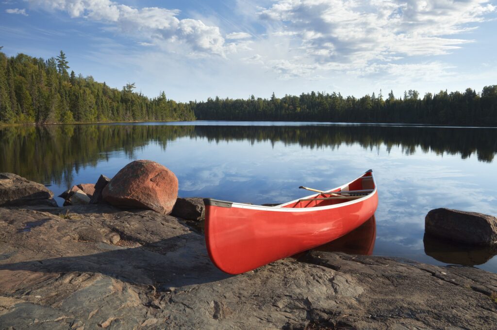 Red canoe on rocky shore of a calm lake surrounded by dense forests under a clear sky