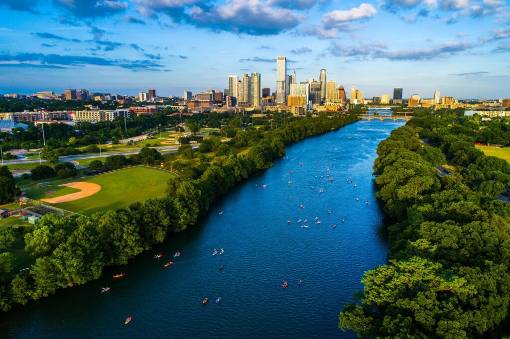 Aerial view of a city skyline with modern buildings beside a river filled with kayaks, surrounded by lush greenery