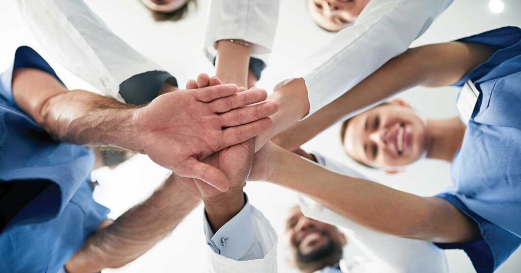 Group of healthcare professionals standing in a circle, hands stacked together in a show of unity and teamwork