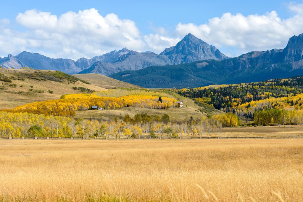 Scenic view of a rural landscape in Colorado with golden autumn foliage, a solitary house, and majestic mountain range in the background