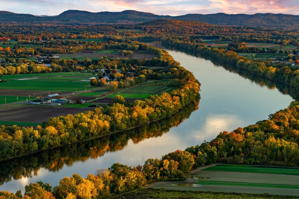 Aerial view of a river winding through lush farmland with colorful autumn foliage and distant mountains