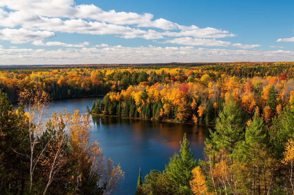 A scenic autumn view of a forest surrounding a calm lake, showcasing vibrant fall foliage under a partly cloudy sky