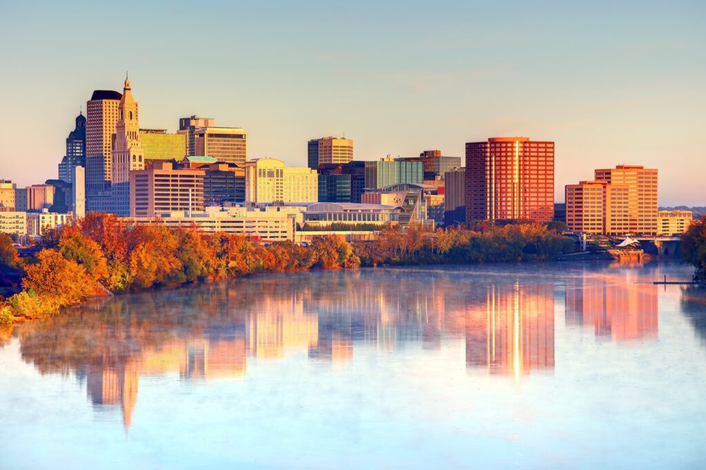 City skyline at sunrise with buildings reflecting on a calm river, surrounded by autumn-colored trees