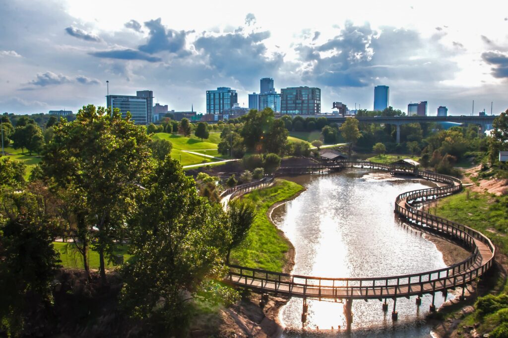 Curved bridge over a reflective pond in a city park, with modern buildings in the background