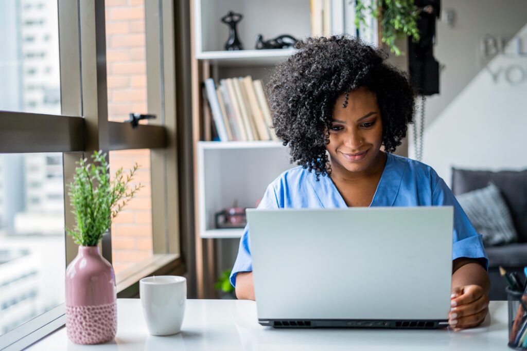 Nurse in blue scrubs working on a laptop in a modern, well-lit home office