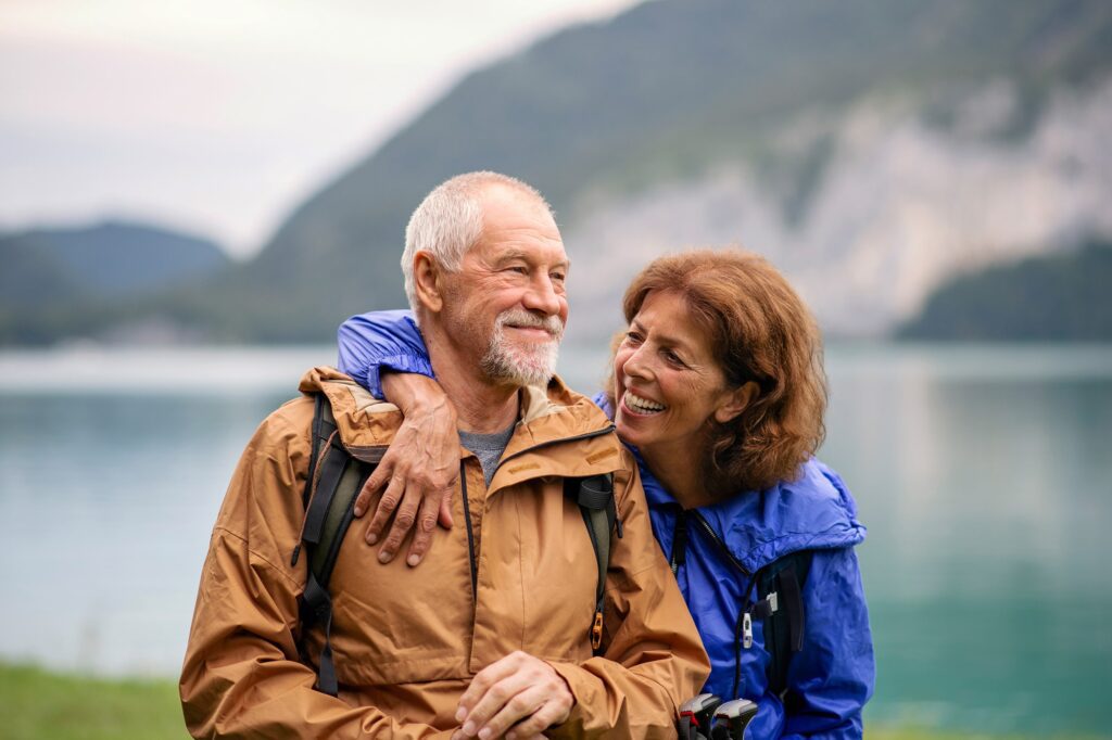 Smiling senior couple in outdoor gear embracing near a lake with mountains in the background