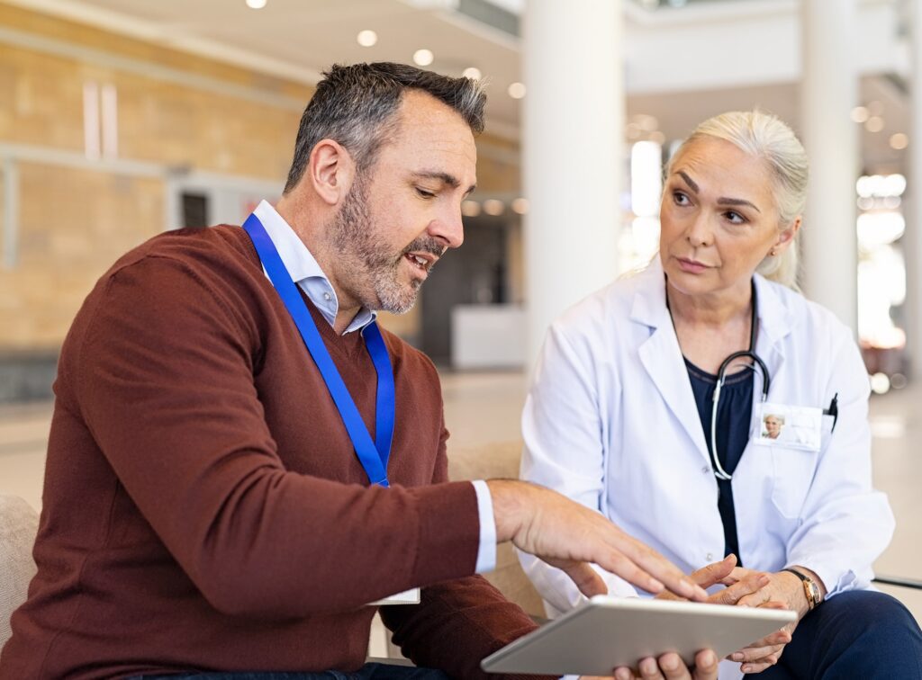 A man in a brown sweater explaining something on a tablet to a female doctor, who is attentively listening