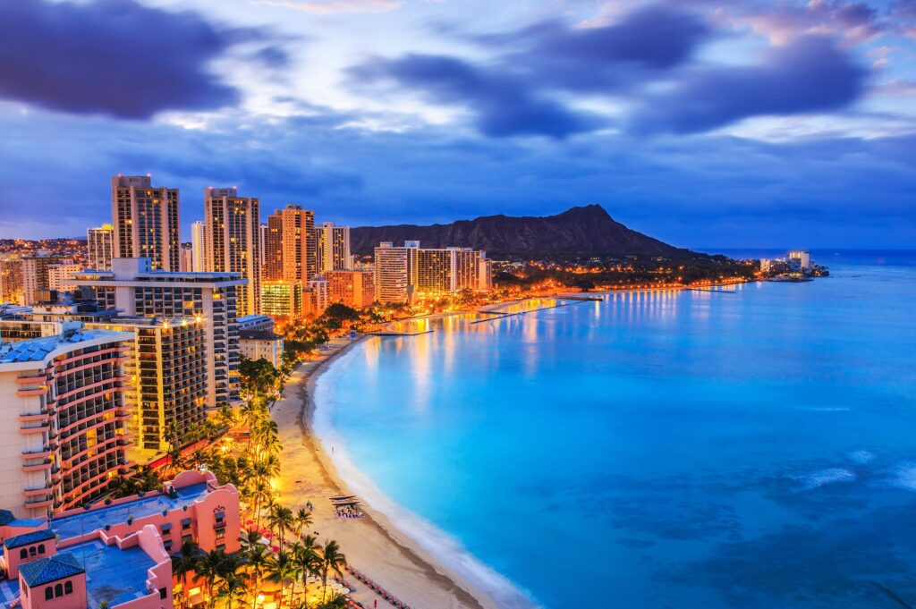 Nighttime cityscape of Honolulu, Hawaii, with bright city lights reflecting on the calm ocean