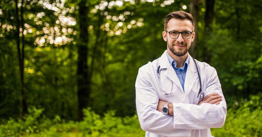 smiling male doctor with arms crossed standing in front of a forest