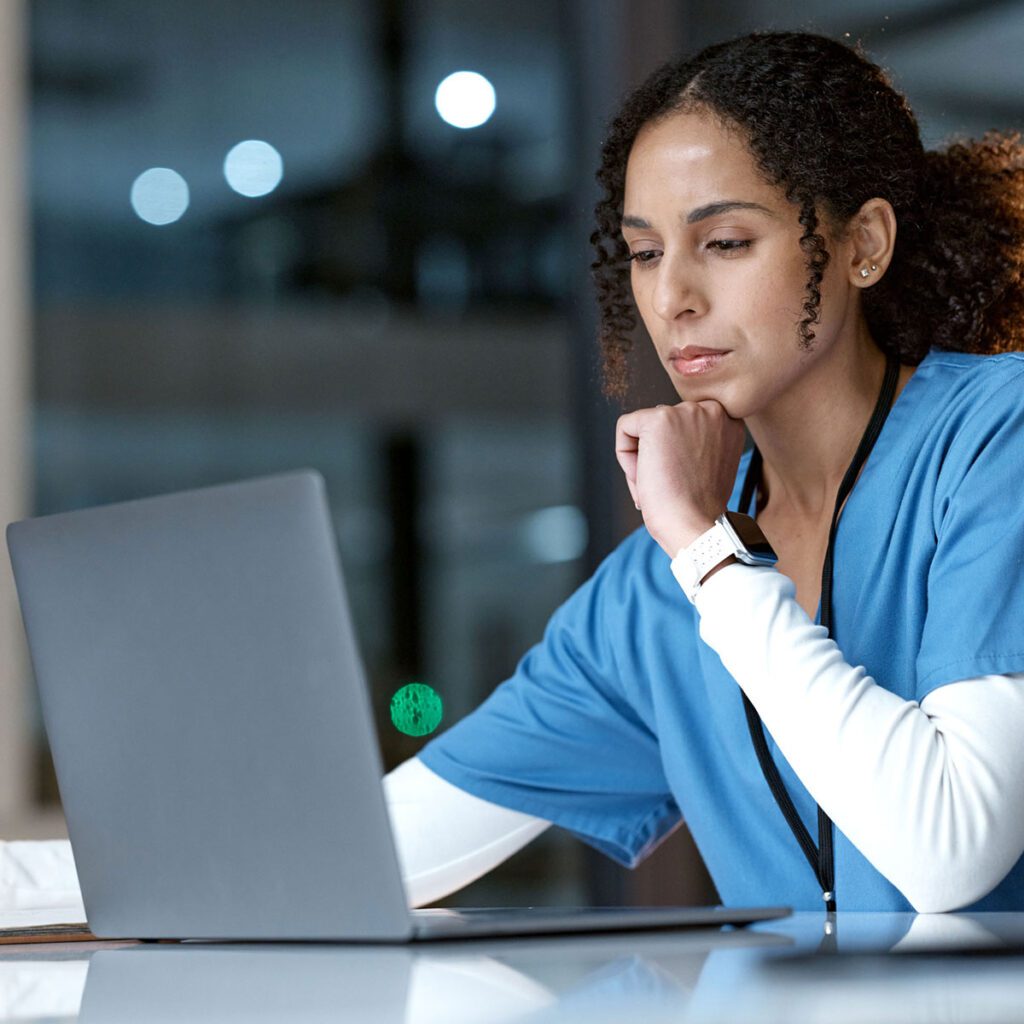 Focused nurse in blue scrubs working on a laptop in a quiet office at night