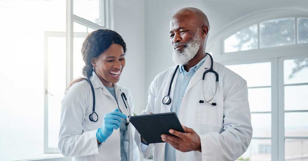 Two doctors in white coats smiling and looking at a tablet