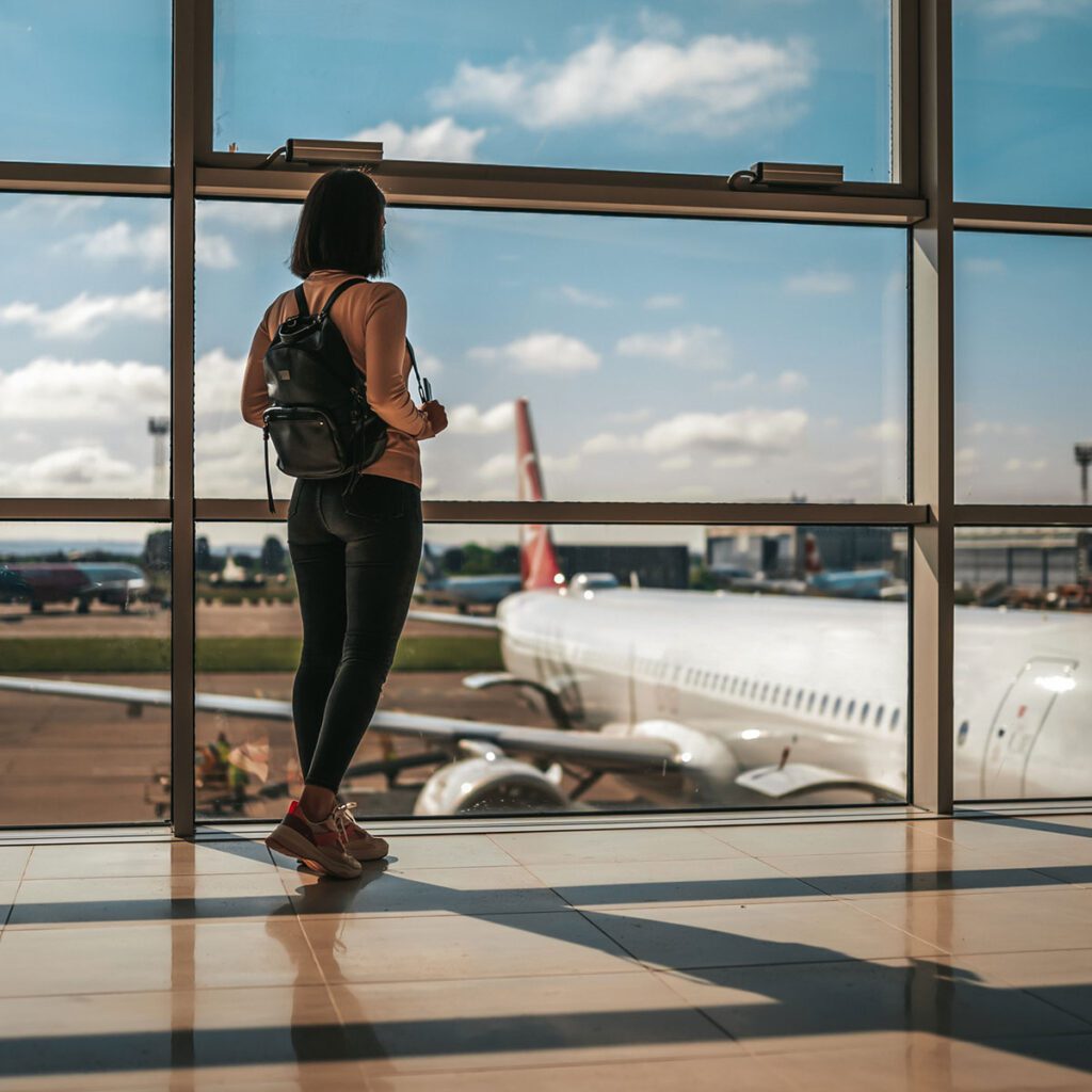 Woman with a backpack standing at an airport window overlooking planes on a sunny day