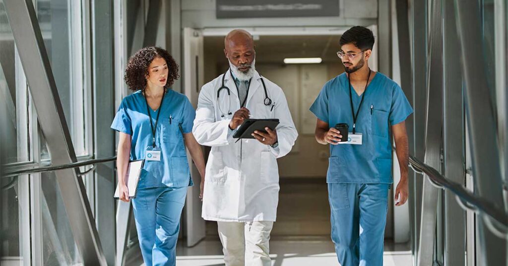 A senior doctor reviews information on a tablet while walking with two younger medical professionals in a hospital corridor