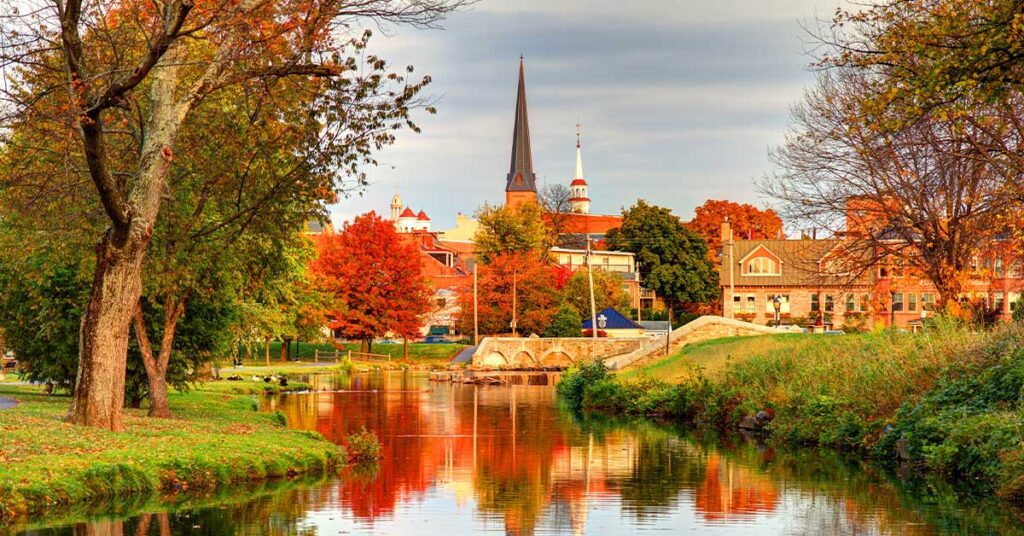 A scenic view of a calm river reflecting the vibrant autumn foliage, with historic buildings and church steeples in the background