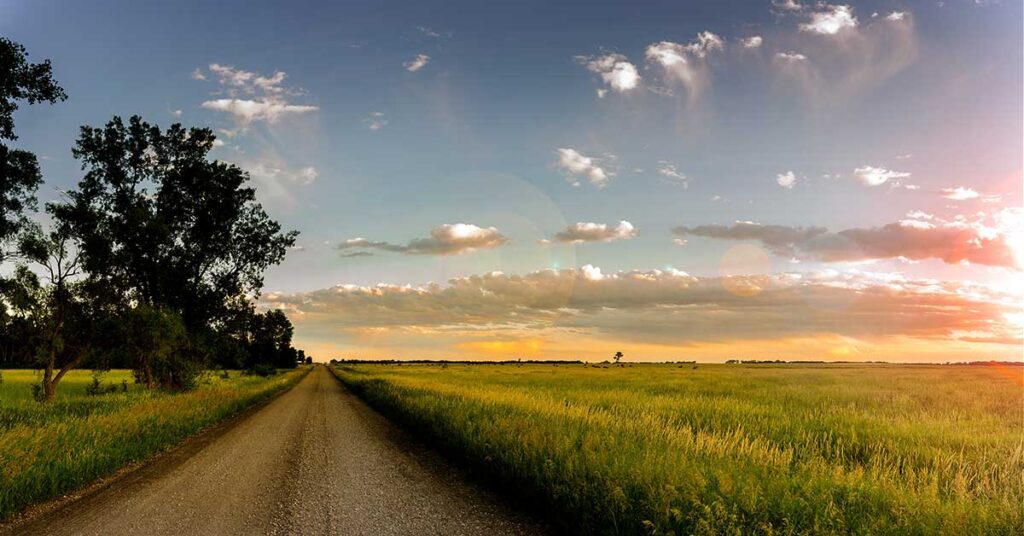 Rural landscape at sunset with a gravel road leading through lush fields under a colorful sky